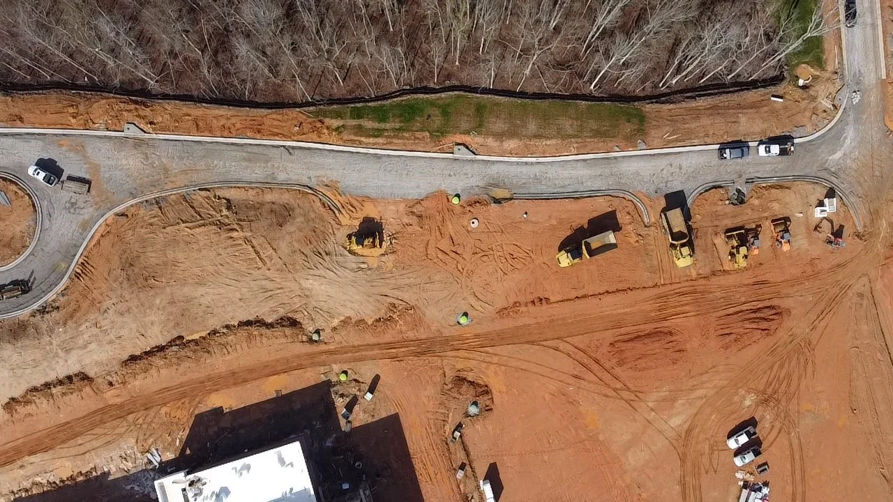 Construction site with dirt and gravel, several construction vehicles, and a partially paved road with some construction work happening.
