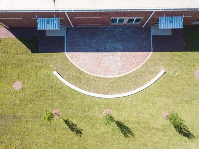 Aerial view of a building with a small curved brick patio, a paved area, and a grassy yard with three trees casting long shadows.
