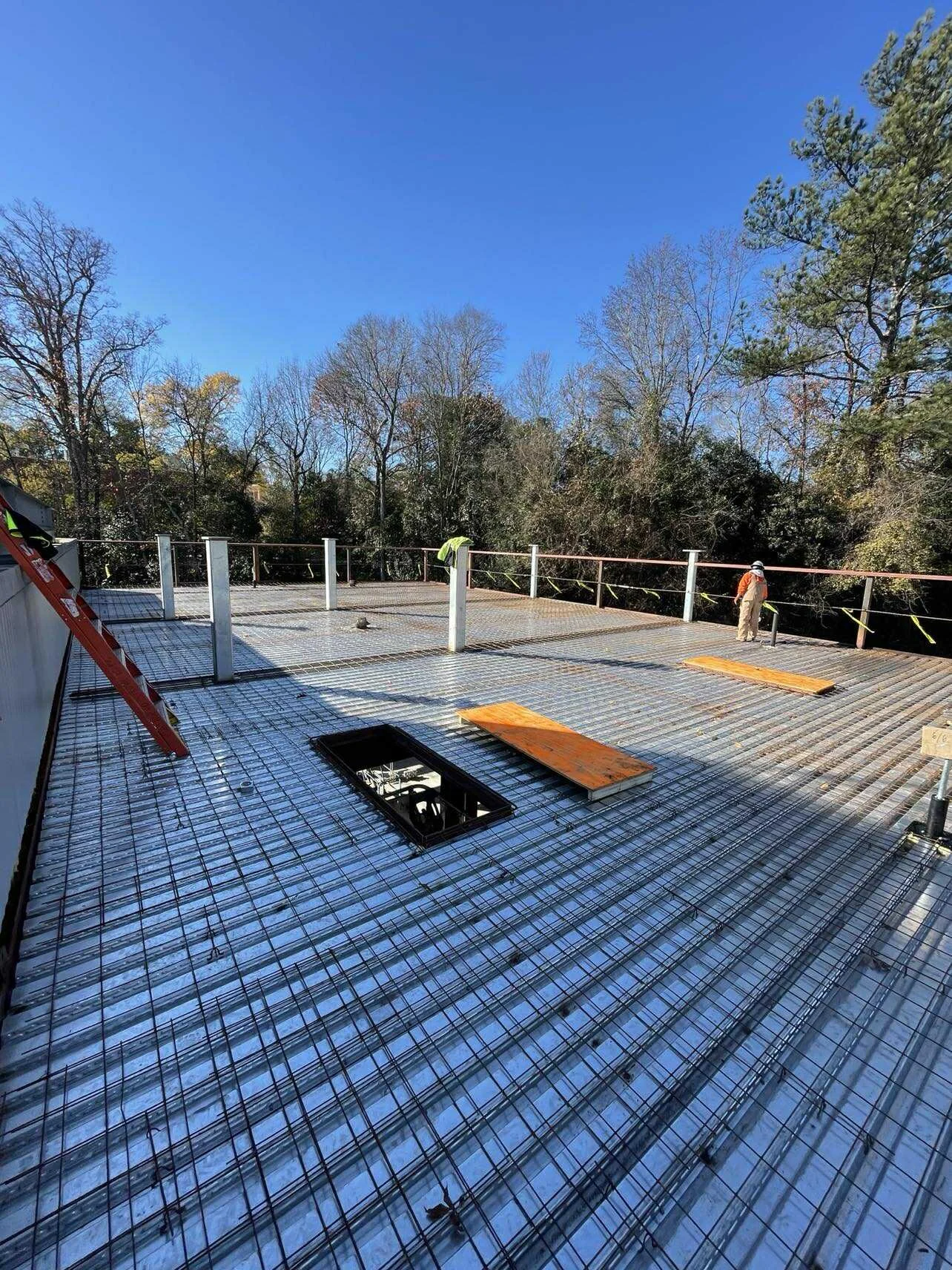 Construction workers installing a metal deck on an elevated building structure with a open hatch and horizontal wooden planks, surrounded by trees under a clear blue sky.