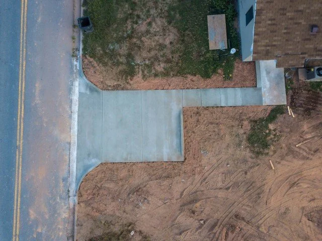An aerial view of a sidewalk and driveway in front of a house, with dirt and grass on either side.