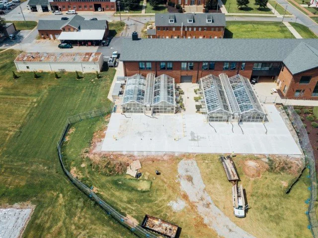 Aerial view of a building with three greenhouses attached to it, surrounded by a grassy yard and some construction materials, in a neighborhood with other residential buildings.