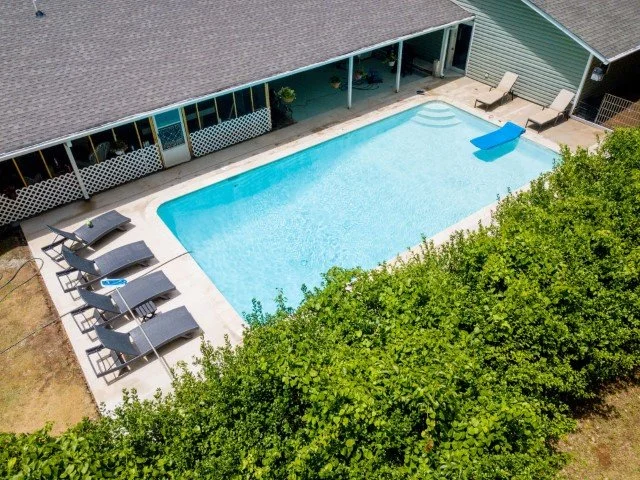 A backyard swimming pool with lounge chairs and a blue float, surrounded by a green hedge and a house with a covered patio area.