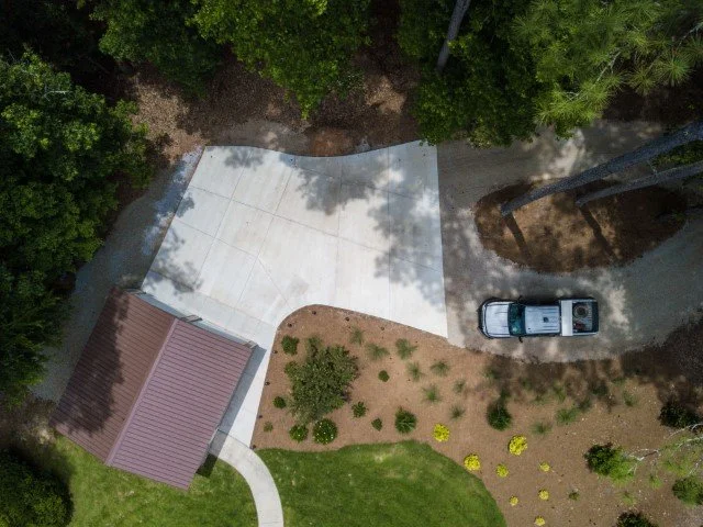 Aerial view of a driveway with a parked truck, adjacent to a small landscaped yard with grass and small bushes, near a house with a brown roof.