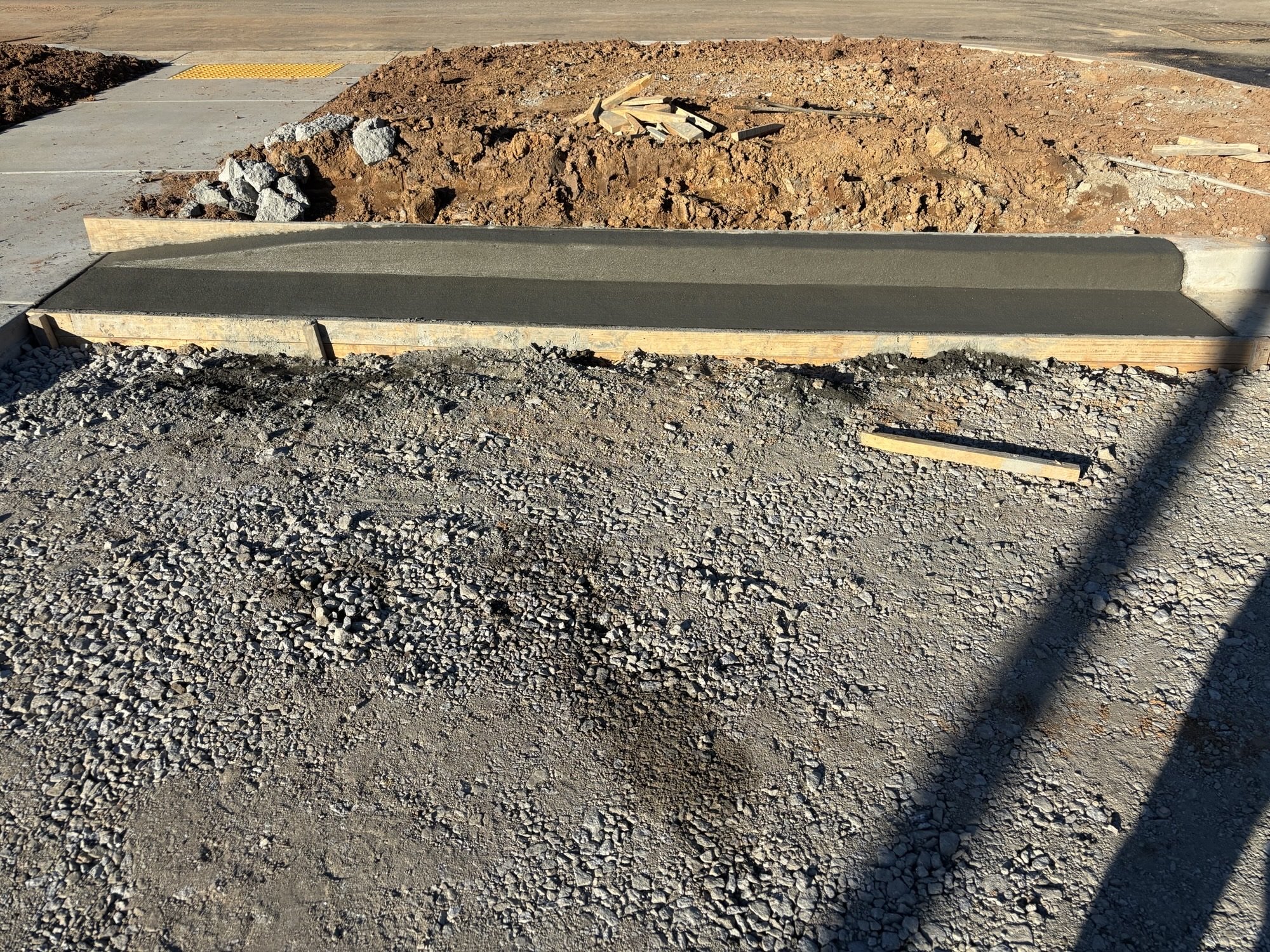 Newly poured concrete sidewalk bordered by gravel and dirt, with a pile of rocks and wooden boards in a construction area.