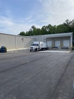 Empty parking lot with a white delivery truck parked near a loading dock at a warehouse, with trees and a blue sky in the background.
