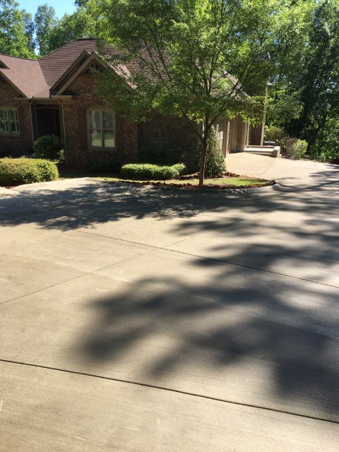 A suburban house with a brick exterior, surrounded by trees and bushes, and a concrete driveway with shadows cast by trees.