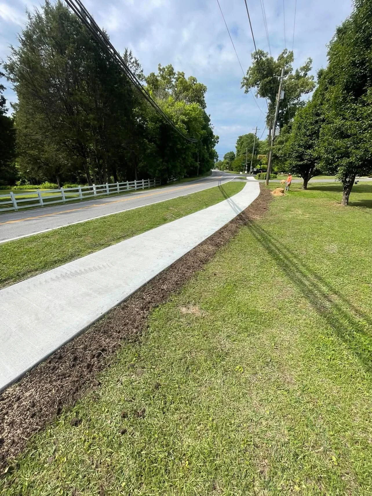 A freshly paved sidewalk runs parallel to a grassy area next to a road. The road has a white fence on one side and is lined with trees. Overhead power lines run alongside the road. A person is sitting on a rock near a tree in the background under a partly cloudy sky.
