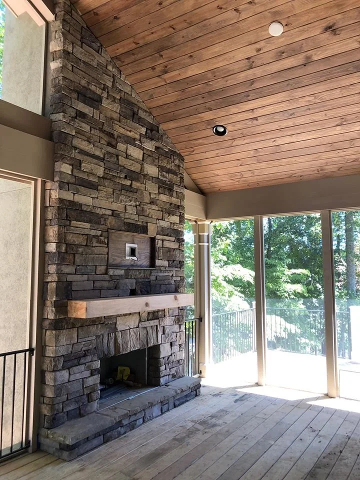 Living room with tall stone fireplace, wooden mantel, wooden ceiling, and large glass windows showing trees outside.