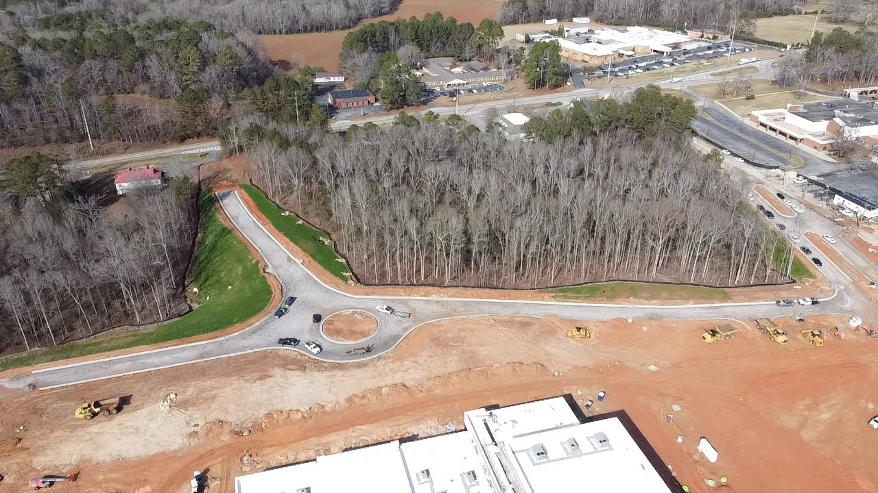 Aerial view of a construction site with a partially built road, a roundabout, trees, and nearby commercial buildings.