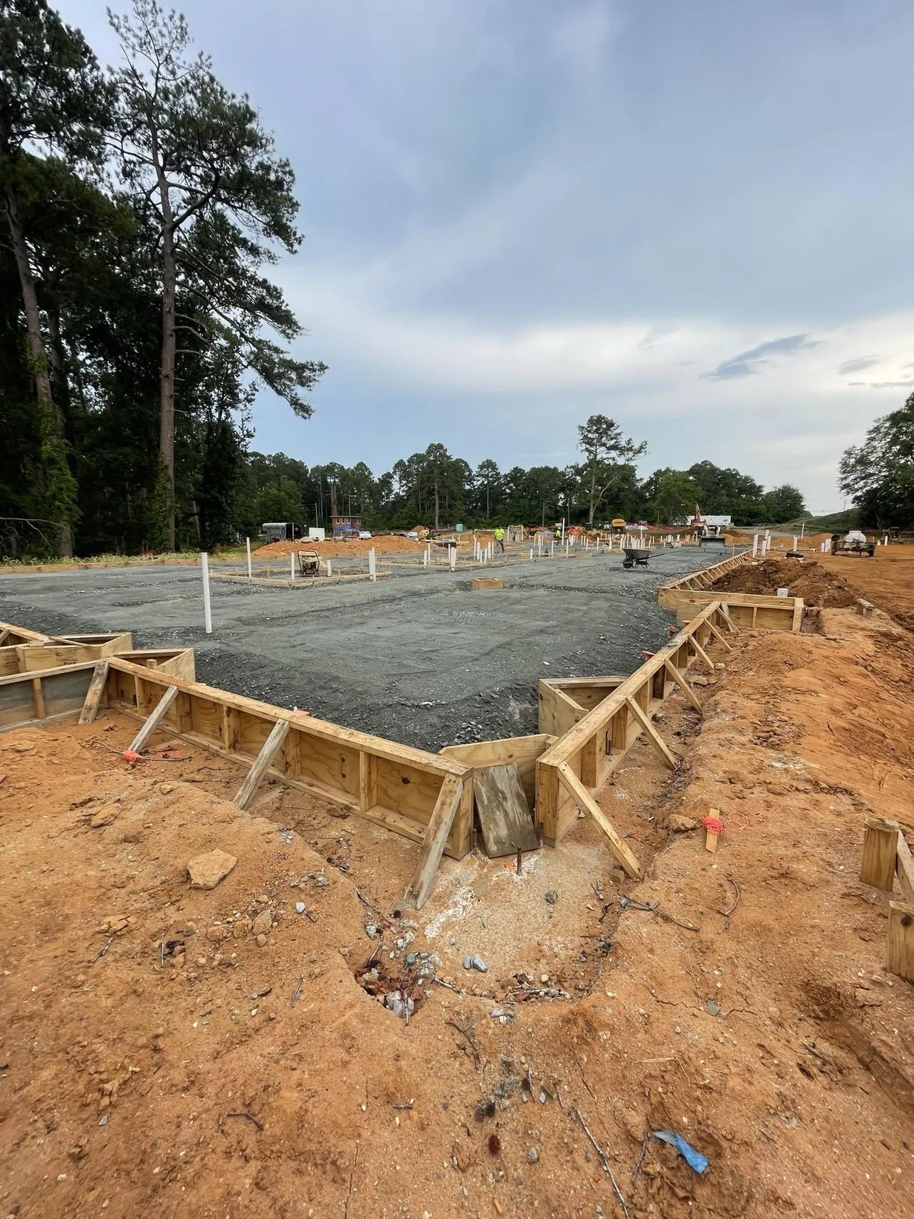 Construction site with wooden framing and gravel foundation for new buildings, surrounded by dirt and trees under a cloudy sky.