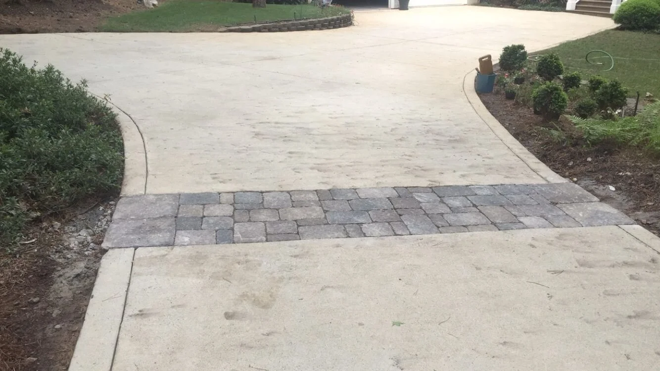 A concrete driveway with a central strip of gray brick pavers, flanked by green bushes on the left and a landscaped garden with plants and garden tools on the right.