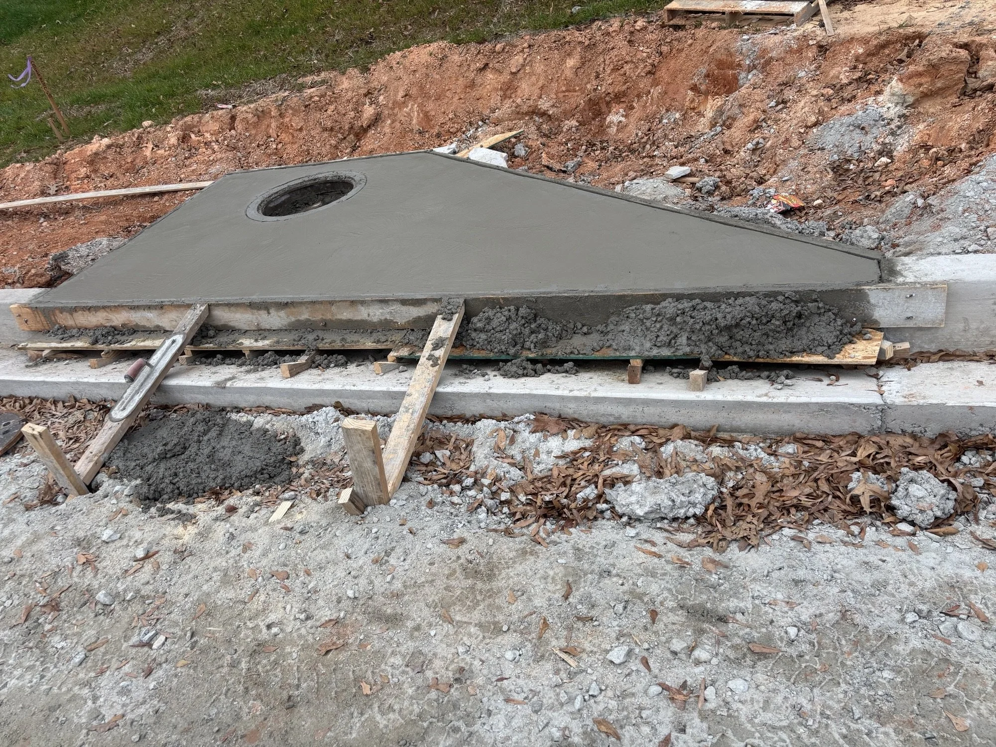 Concrete sidewalk being poured and formed in a construction site, with wooden forms and a hole in the concrete for a drain or utility access.