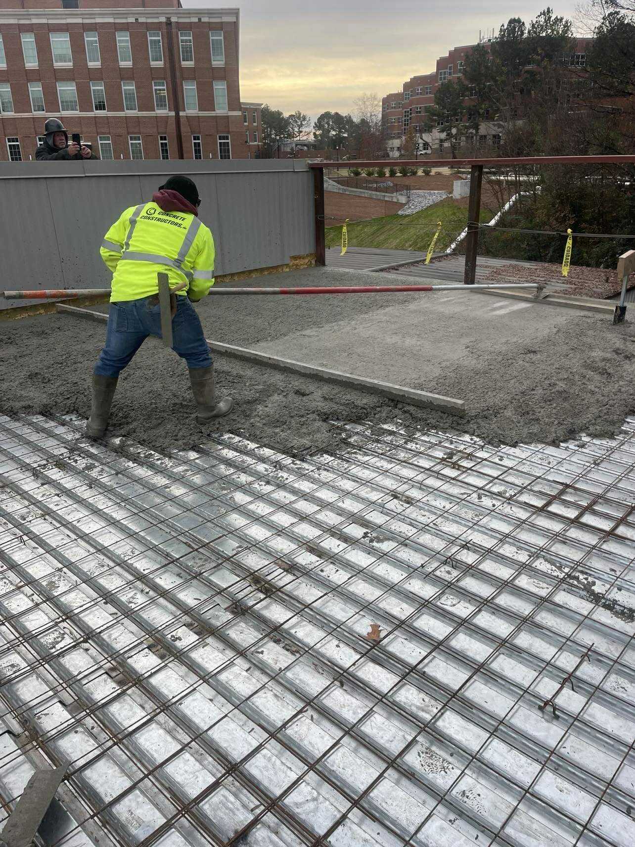 Construction worker smoothing wet concrete on a balcony with rebar grid at sunrise, with urban buildings and trees in the background.