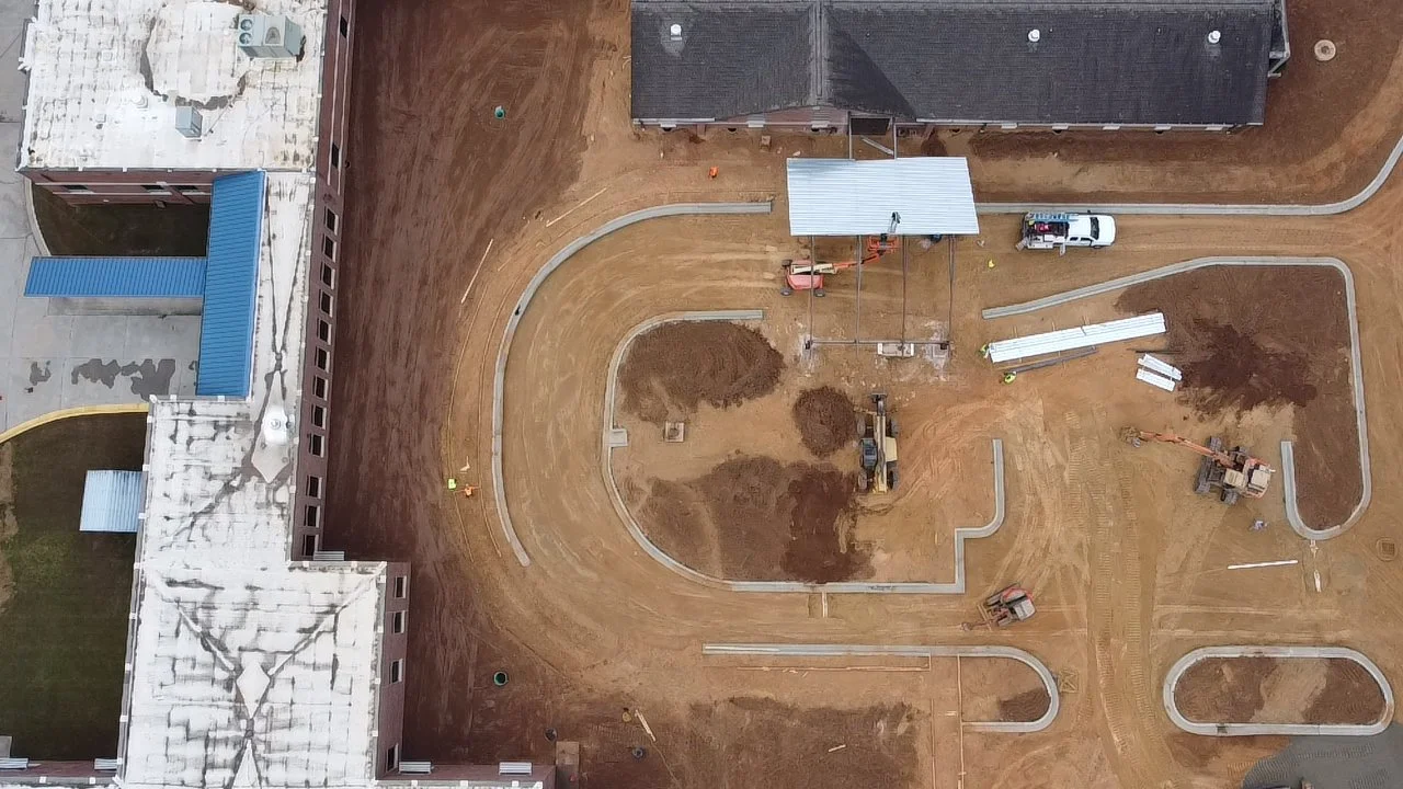 An aerial view of a construction site where a new pathway or sidewalk is being built within a residential area. Several construction vehicles and workers are present, and some parts of the ground are still dirt with piles of soil.