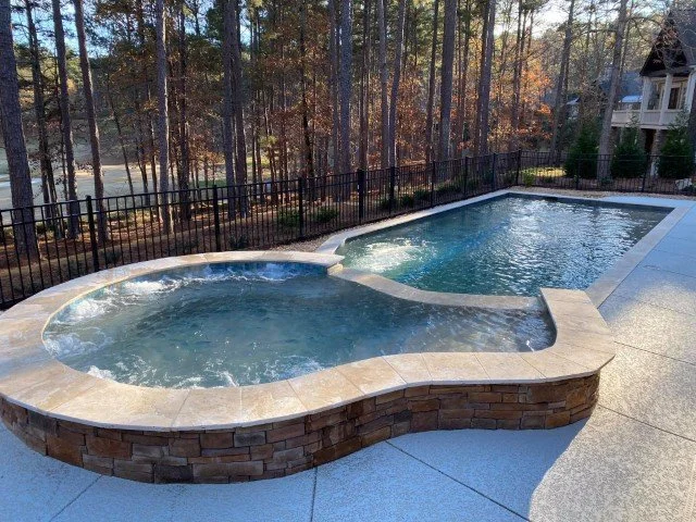 A backyard pool with a built-in hot tub, enclosed by a black metal fence, set against a wooded area with tall trees.