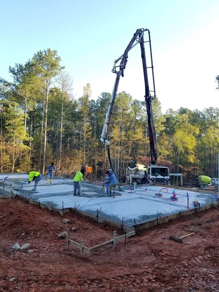 Construction workers are pouring concrete on a building foundation with a concrete pump truck, on a clear day surrounded by trees.