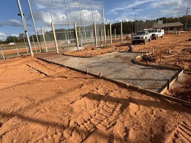 Under construction baseball field with a concrete slab, wooden forms, and equipment, with parked trucks nearby.