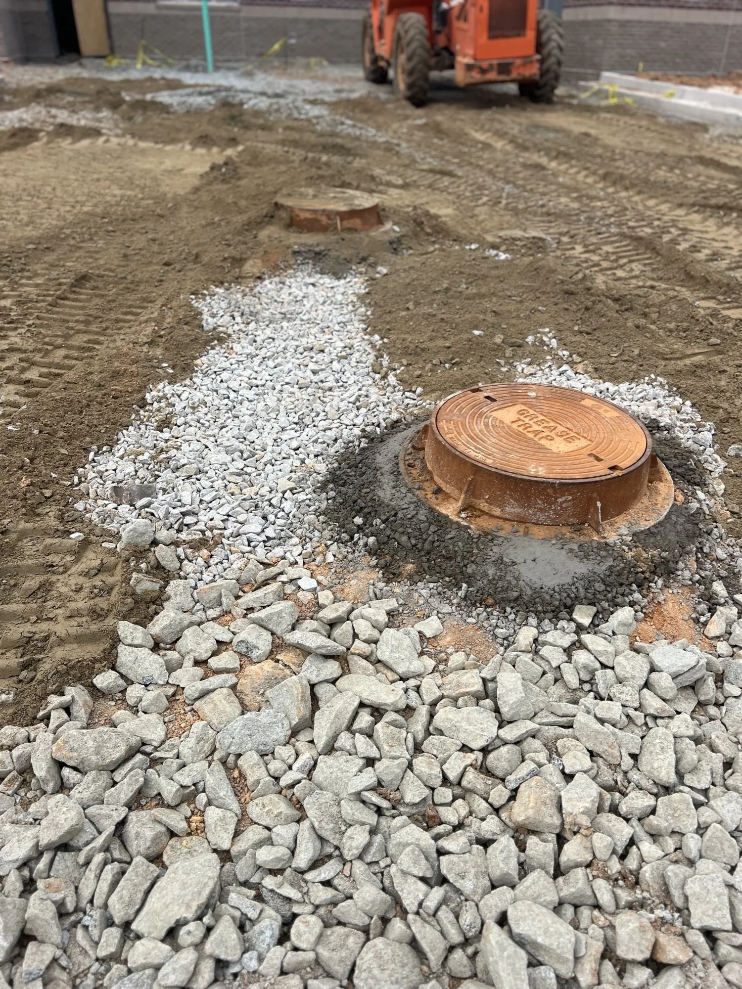 Construction site with gravel and dirt, orange manhole covers, and an orange construction vehicle in the background.