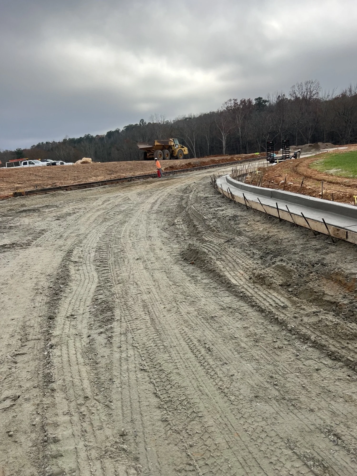 Construction site with dirt road, a worker wearing safety orange vest, a large yellow dump truck, and construction trailers, with trees and overcast sky in the background.