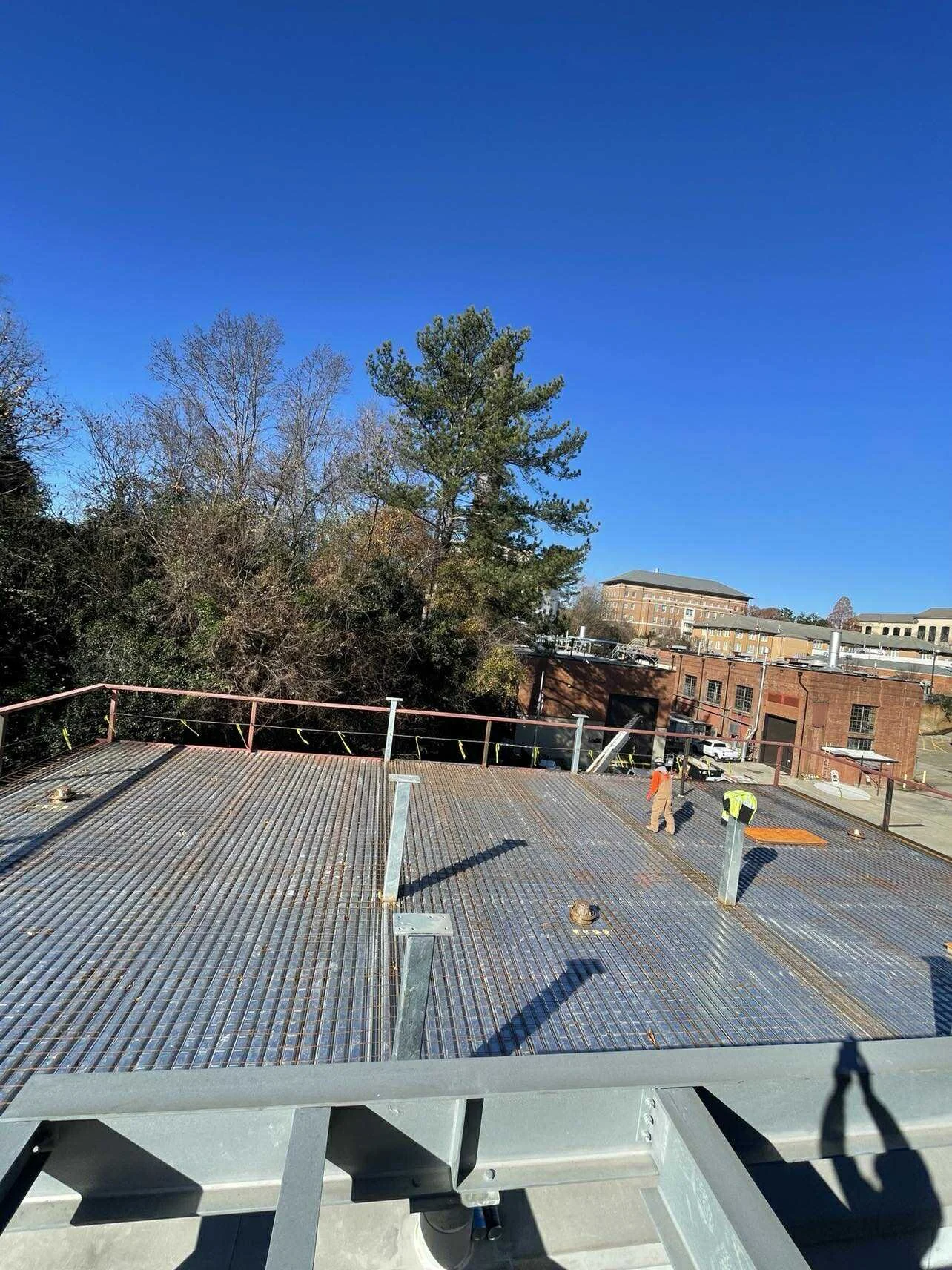 Construction workers on a metal rooftop under a clear blue sky with trees and buildings in the background.