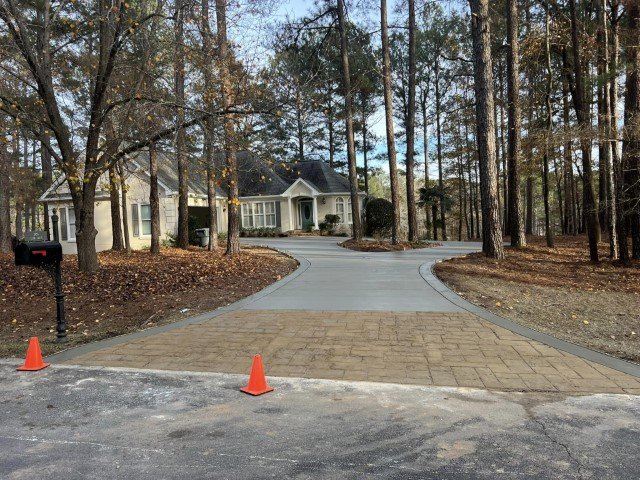 Concrete driveway leading to a house with a landscaped yard surrounded by trees, with two orange traffic cones and a mailbox on the side.