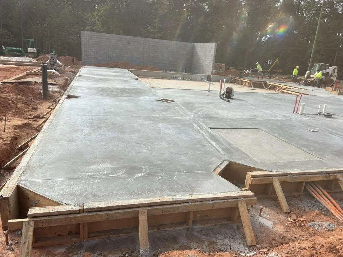 Construction site with a partially completed concrete foundation, wooden forms, and construction workers at work in the background.