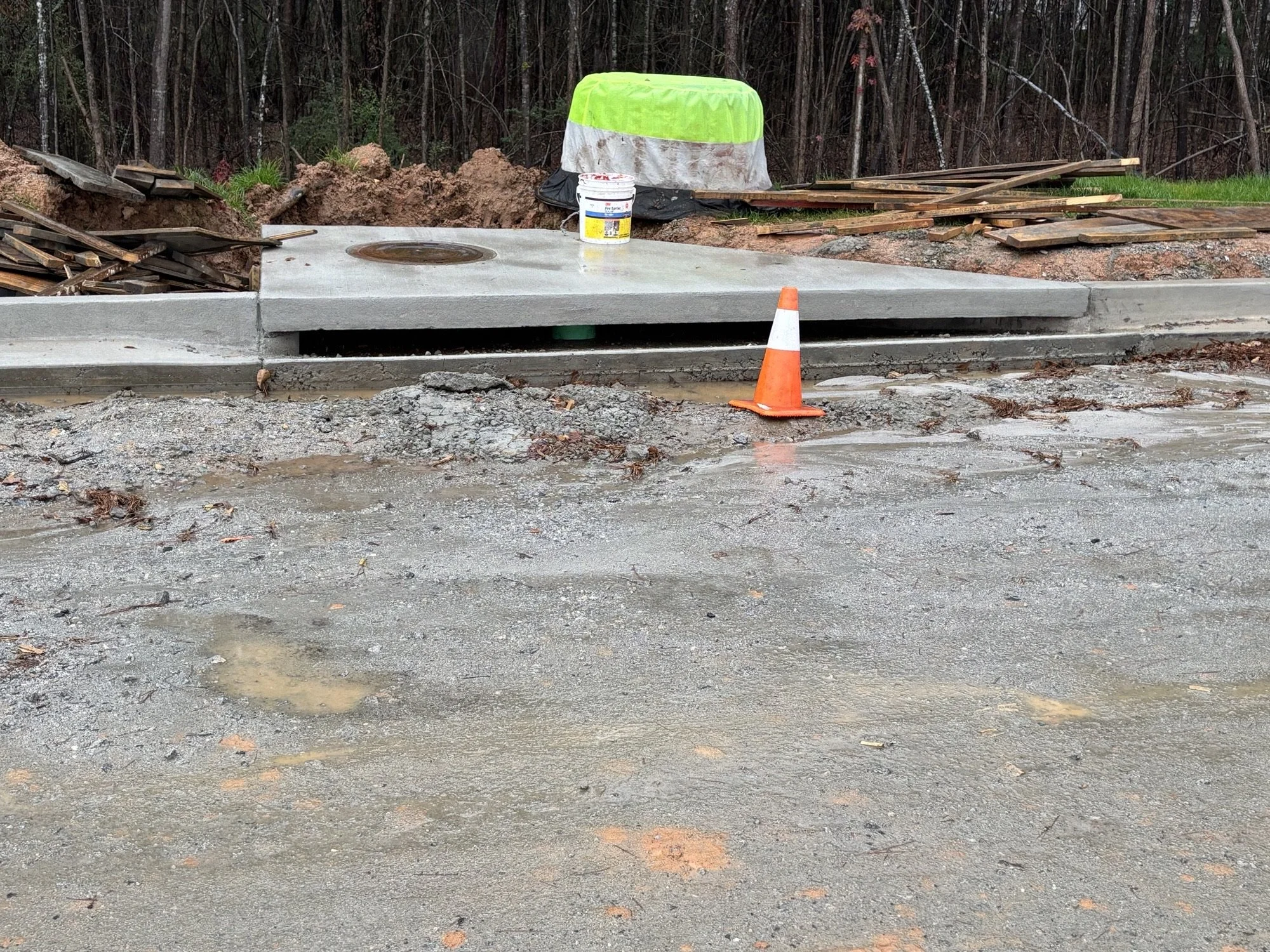 Construction site with a freshly poured concrete sidewalk, orange safety cone, and construction materials including wood planks and a bucket, with a wooded background.