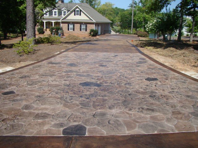 View of a newly paved stone driveway leading to a house with a porch, surrounded by landscaping and trees.