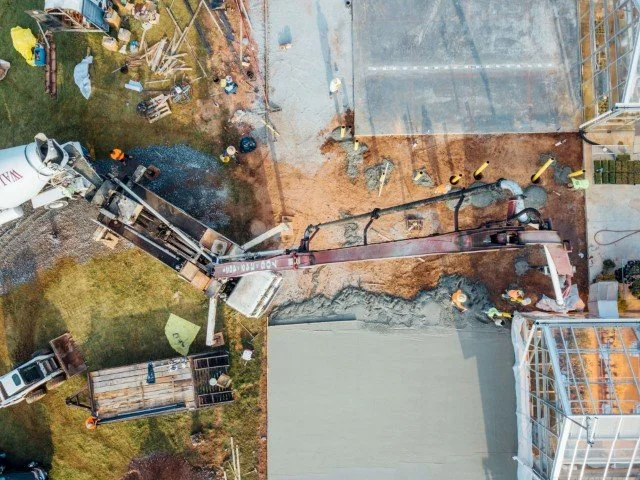 An aerial view of a construction site with a concrete pump truck pouring concrete into a foundation. Construction workers are present, and a partially built structure with scaffolding can be seen on the right.