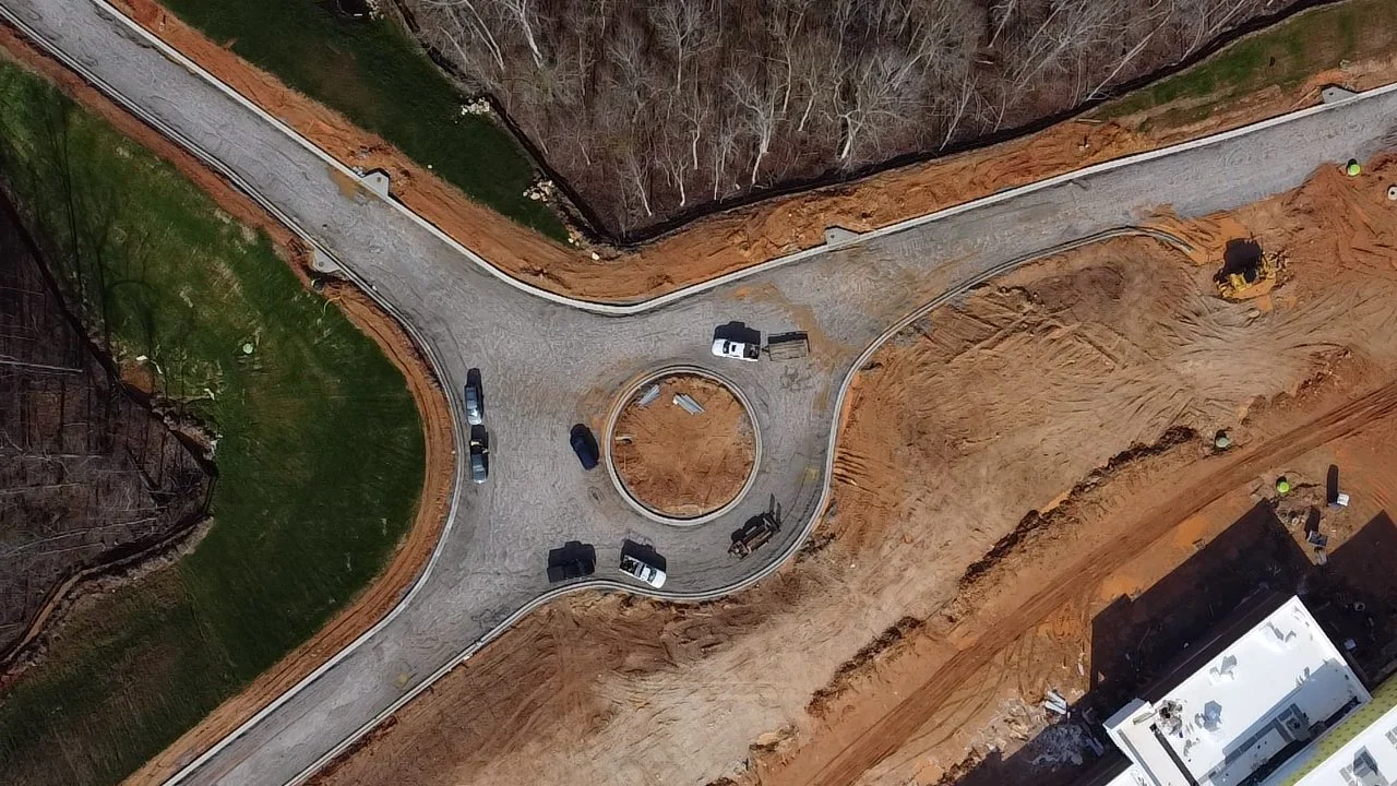 An aerial view of a construction site showing newly paved circular driveway with surrounding gravel and dirt, some construction vehicles parked, and a section of grass and trees.