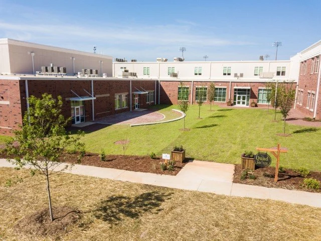 A courtyard with a green lawn, young trees, a concrete pathway, and a sign that says 'Quad' in front of a building with white and brick walls under a blue sky.