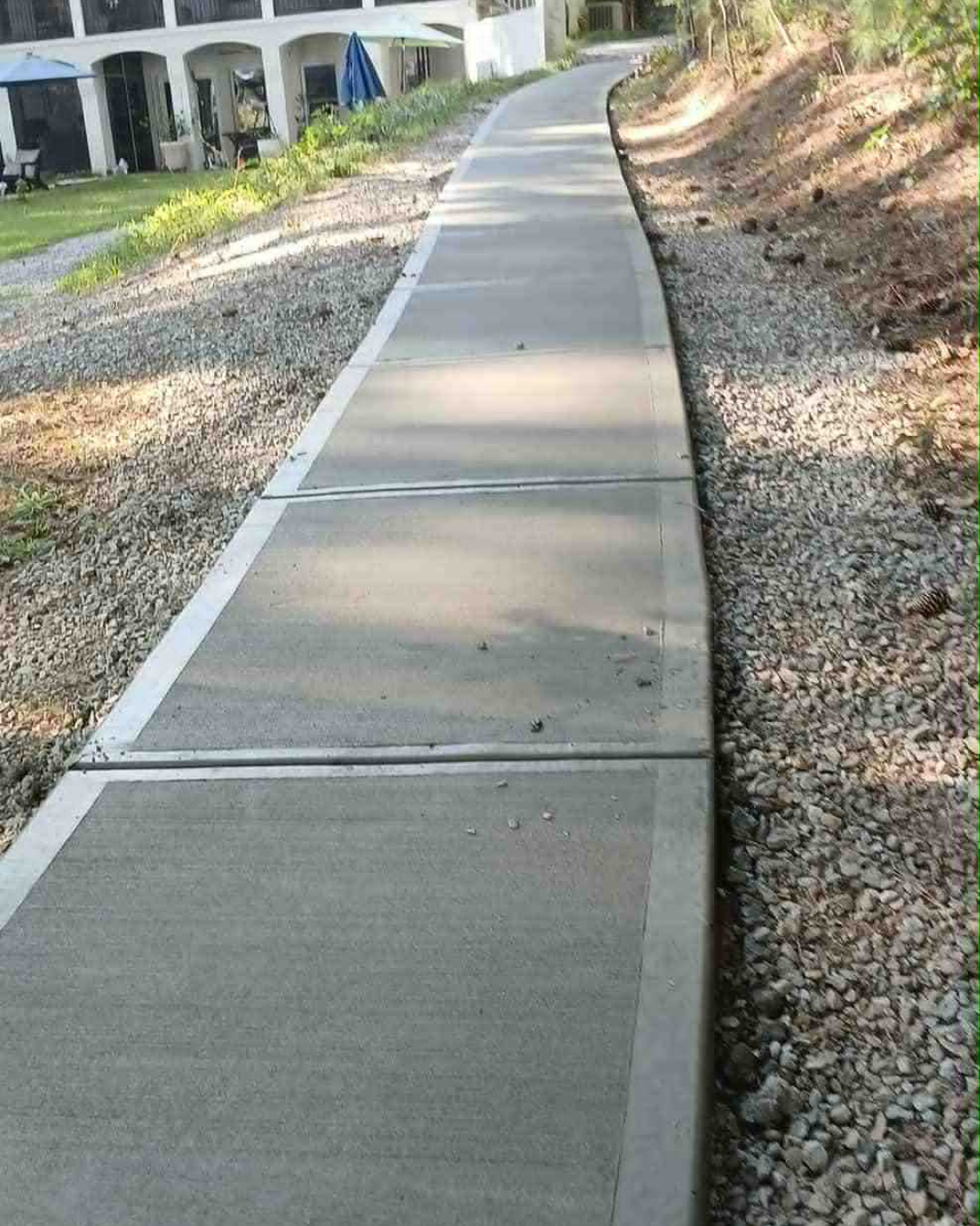 A curved concrete sidewalk next to a gravel path with a grassy area and a building with balconies in the background.