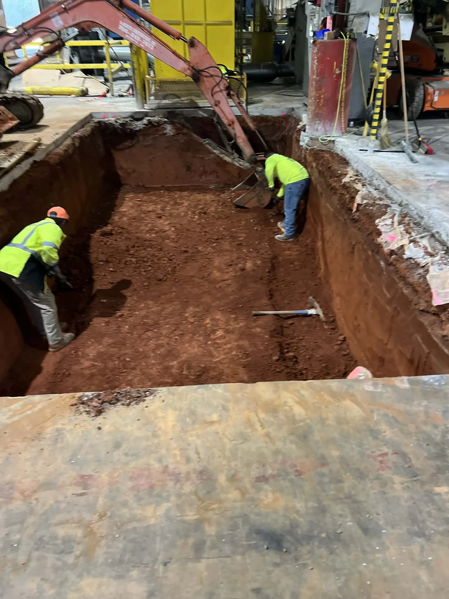 Construction workers with safety equipment working in a large excavation site with heavy machinery and industrial equipment surrounding them.