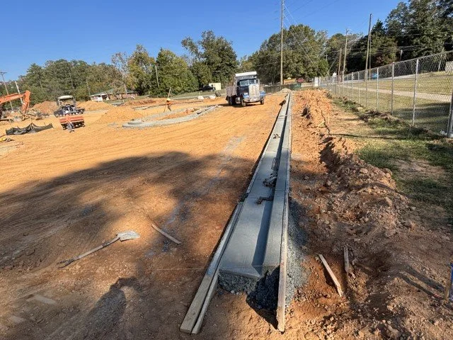Construction site with a partially installed metal guardrail on a dirt road, construction vehicles, and equipment in the background, surrounded by trees and a fence.