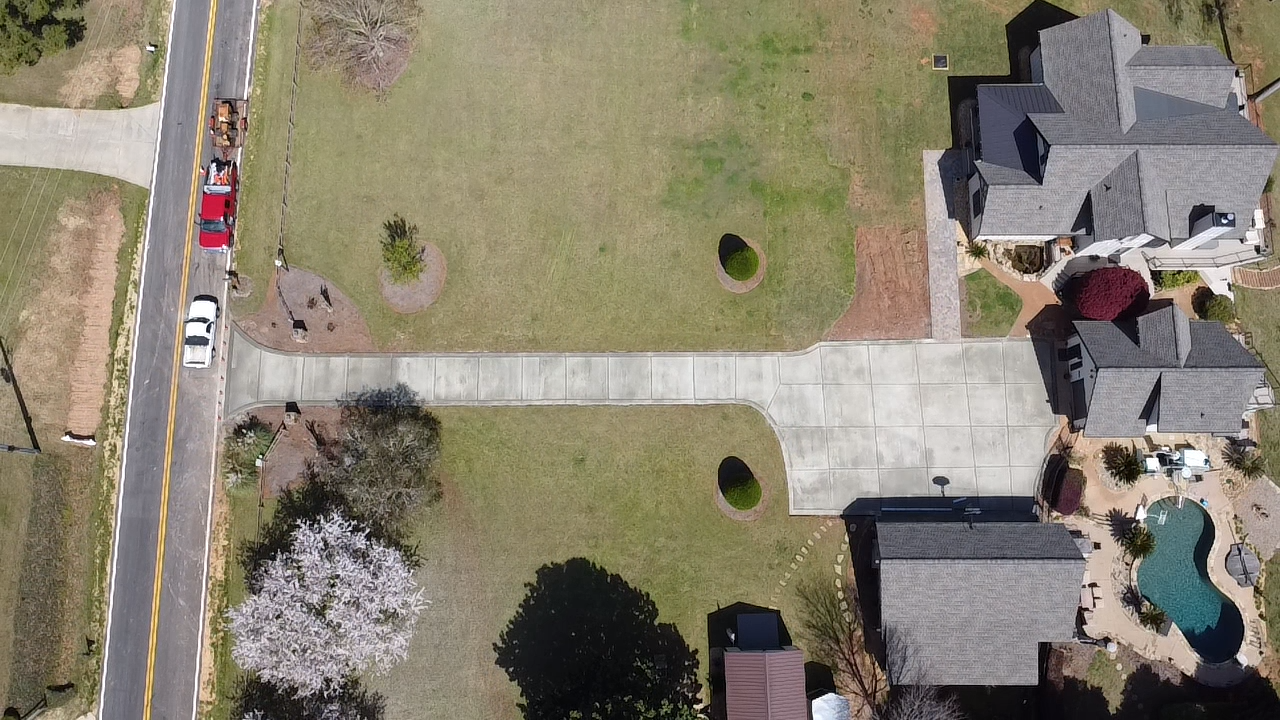 Aerial view of a residential neighborhood showing a house with a pool, a driveway, and a lawn area with trees and bushes.