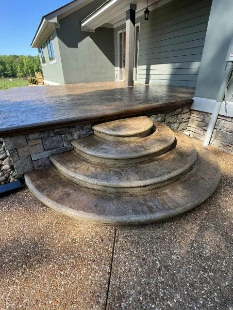 Outdoor steps leading from a patio to a house entrance, with three rounded stone steps and a landing, beside a gray house with siding and a garage door.