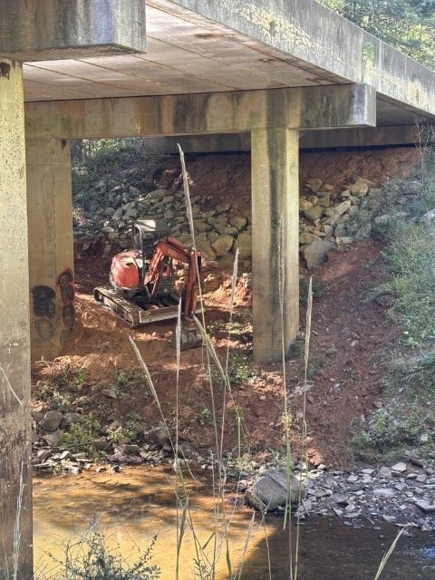 Under a concrete bridge over a small river, a mini excavator with a red body and tracks is positioned on the muddy riverbank.
