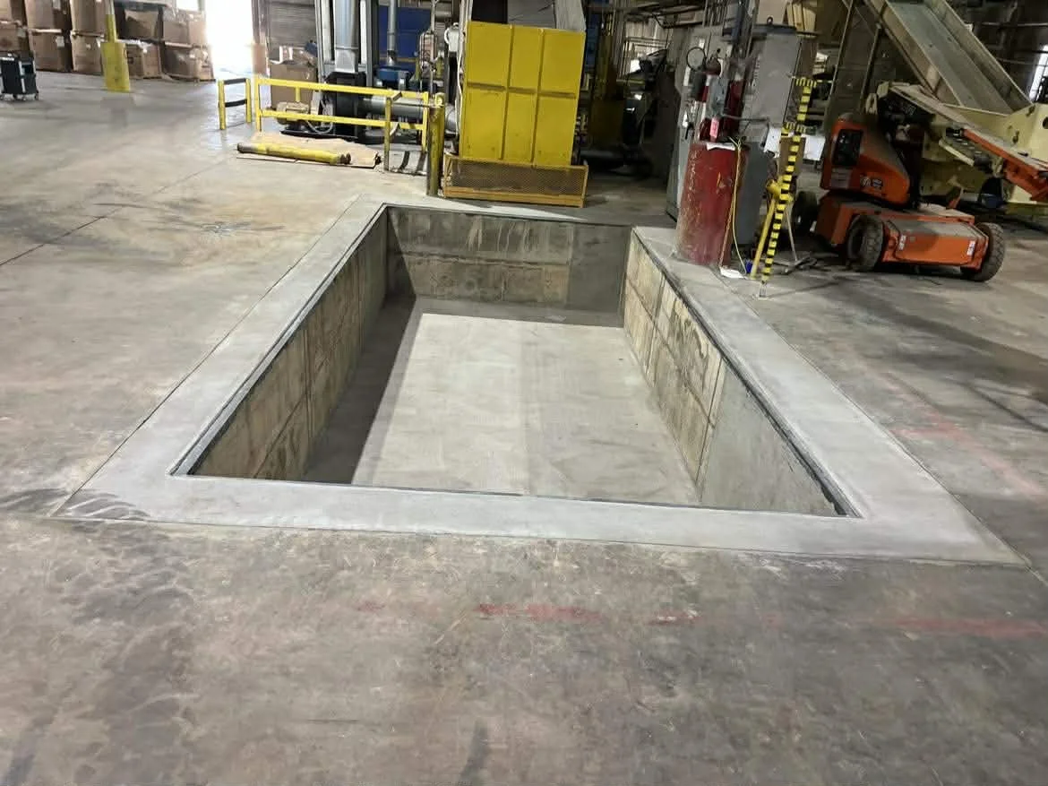A rectangular hole or pit in the concrete floor of an industrial or warehouse setting, with surrounding concrete borders and construction equipment and safety barriers in the background.
