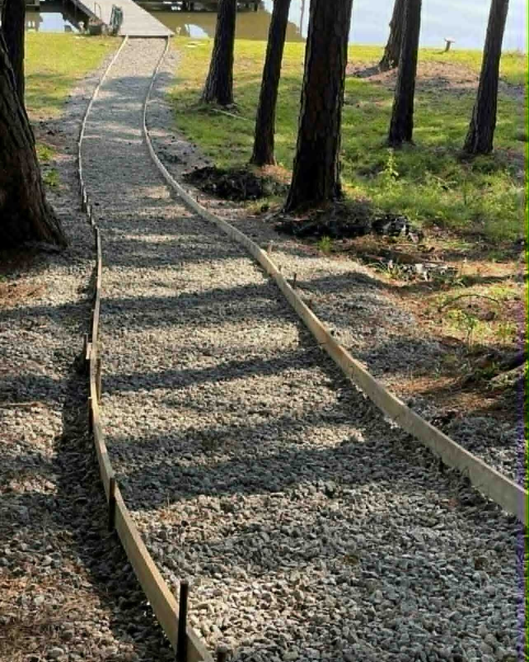 A gravel pathway under construction in a wooded area, with low wooden boards outlining the sides, leading towards a body of water in the background, surrounded by trees and grass.