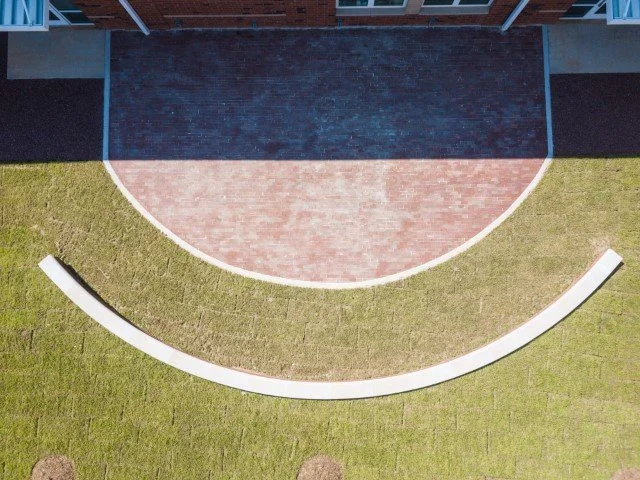 Aerial view of a small outdoor amphitheater with a curved brick stage, surrounded by a grassy area and concrete walking paths.