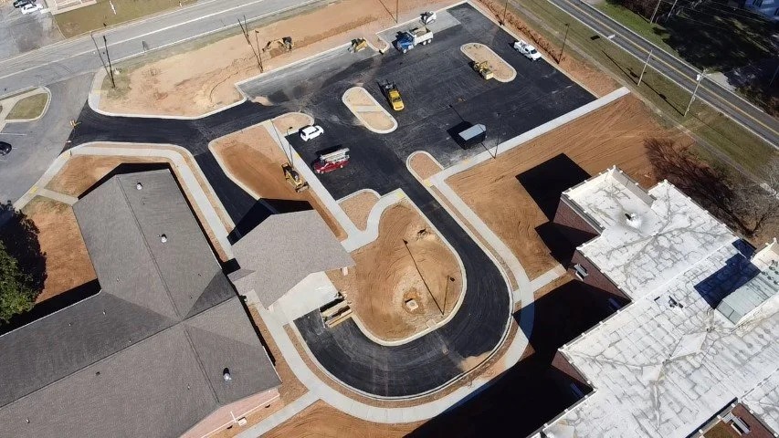 A new, paved parking lot under construction with marked parking spaces, curbs, and sidewalks, next to residential buildings, with construction equipment and vehicles present.