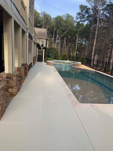 A backyard pool area with a concrete walkway, a swimming pool with an attached hot tub, and surrounded by trees and a black fence.