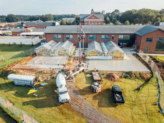 Construction site with concrete being poured for a patio area, greenhouses, and building in the background.