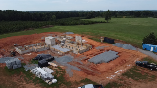 Construction site with foundation work, construction materials, and equipment on red dirt, surrounded by green fields and trees.