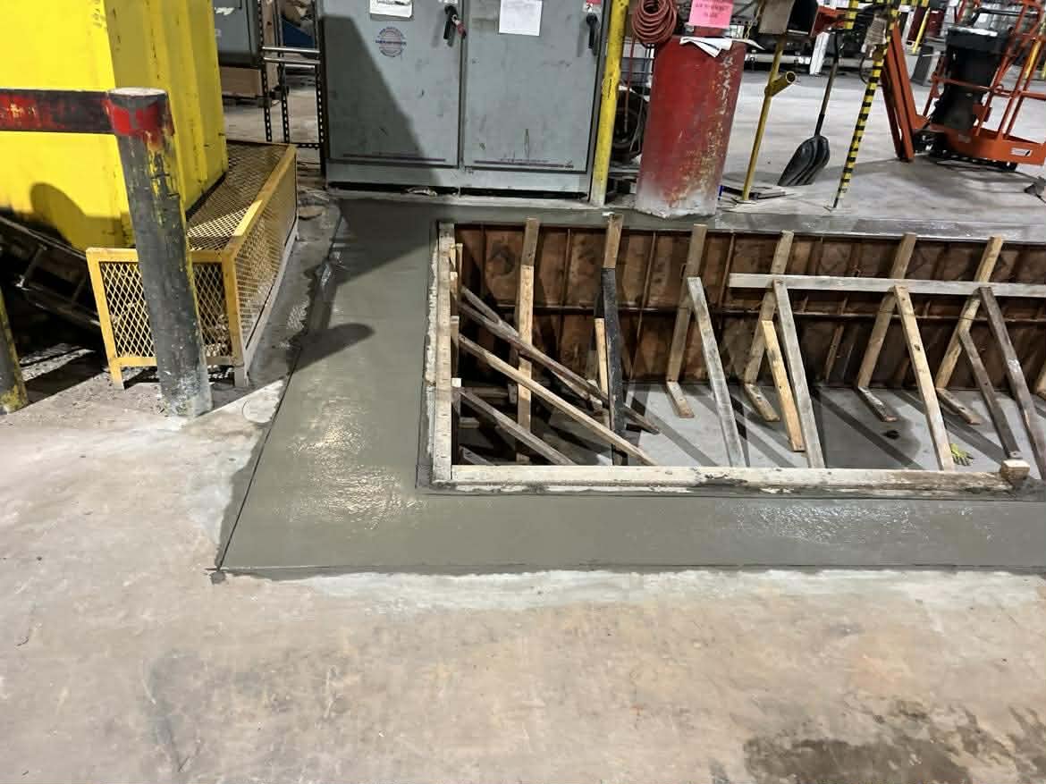 A construction area with an opening in the floor reinforced with wooden beams, surrounded by fresh concrete and construction tools and equipment nearby.