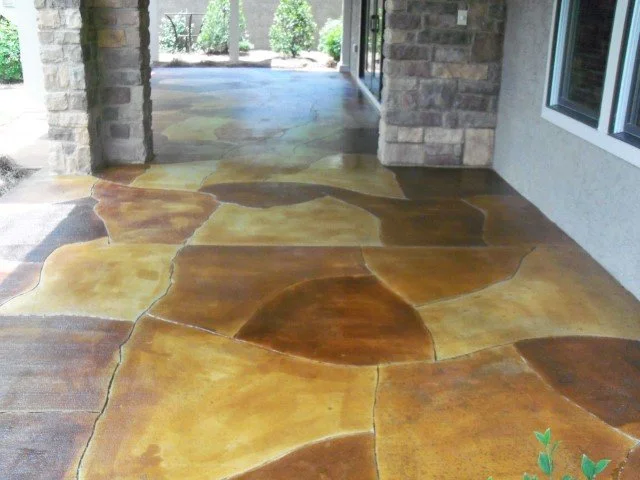 Colorful stone patio with irregularly shaped stones in shades of yellow, orange, and brown, adjacent to a brick wall and large window, with a view of green trees and shrubs in the background.