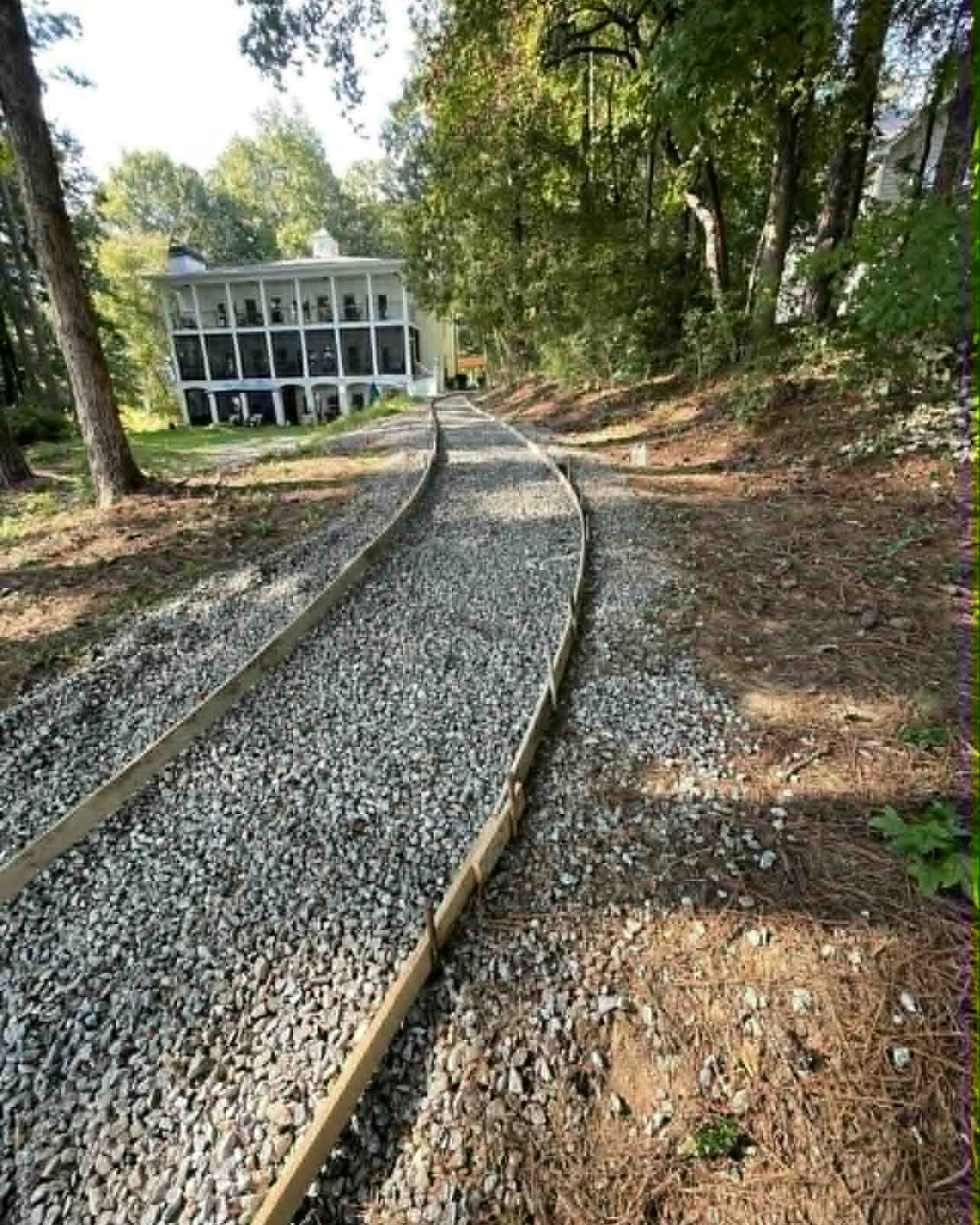 A narrow railroad track on a gravel path surrounded by trees, leading up to a white house with large windows on a porch, situated on a grassy area.
