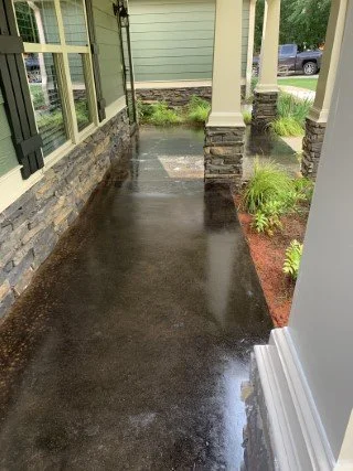 Wet concrete porch with brick wall, window, and plants, next to a house with columns and a driveway in the background.