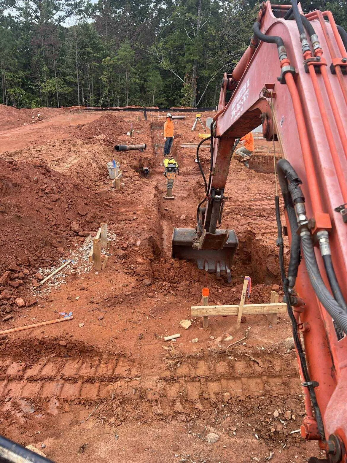 Construction workers operating an excavator and working on a dirt site surrounded by trees.