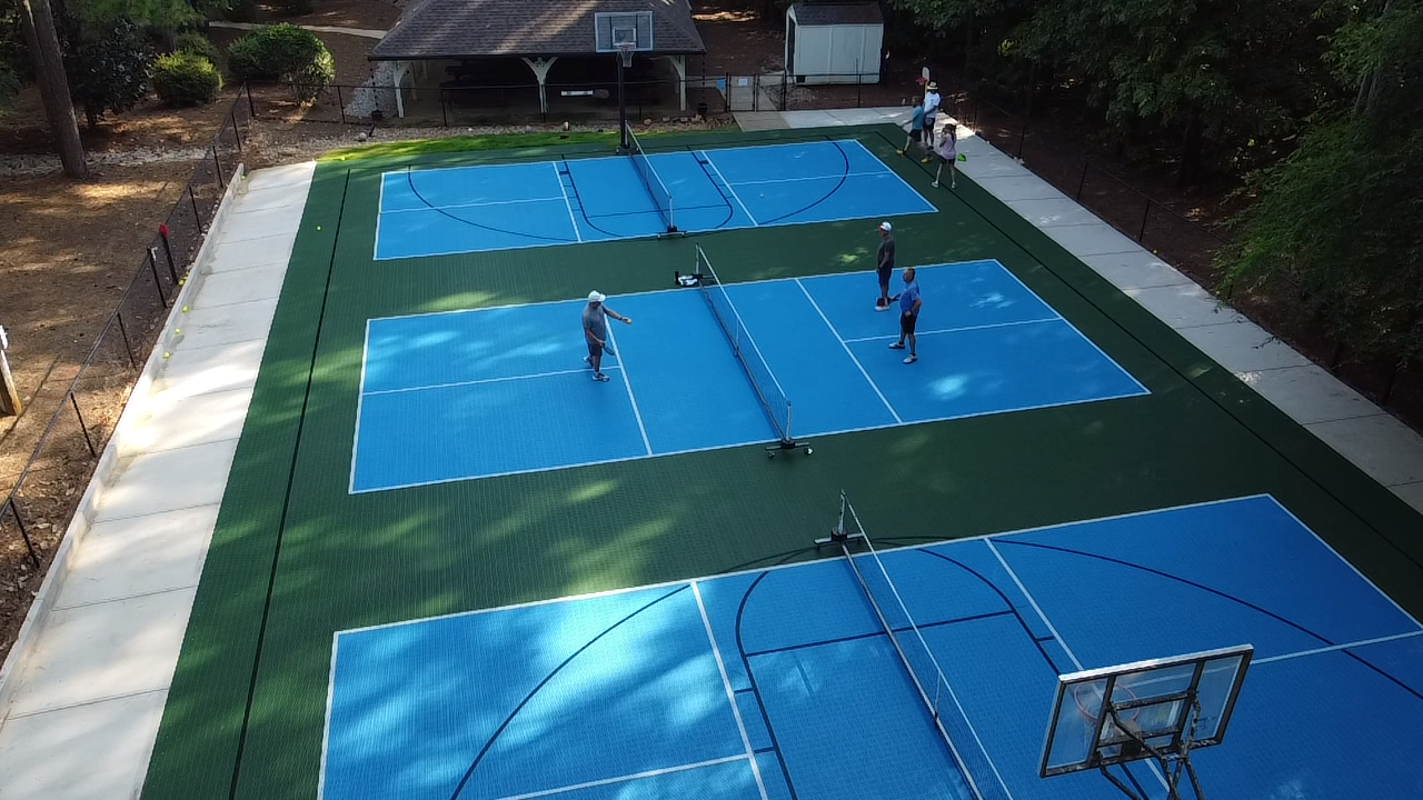 An aerial view of a multi-court basketball and pickleball facility with people playing and walking around, surrounded by trees and a fence.